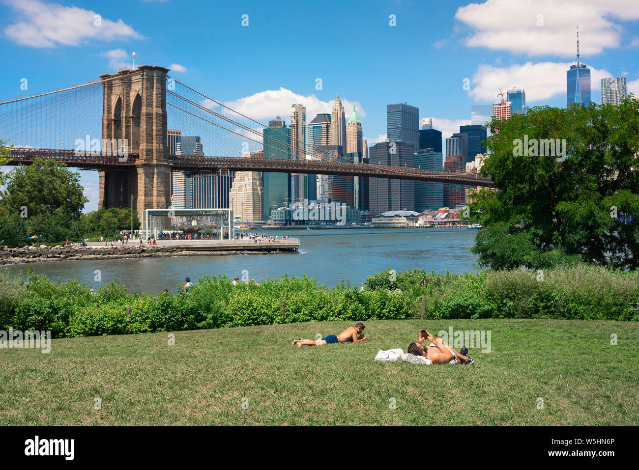 New York city summer, view of two men sunbathing in Main Street Park, Brooklyn, with the Brooklyn Bridge and Lower Manhattan skyline in the distance. Stock Photo