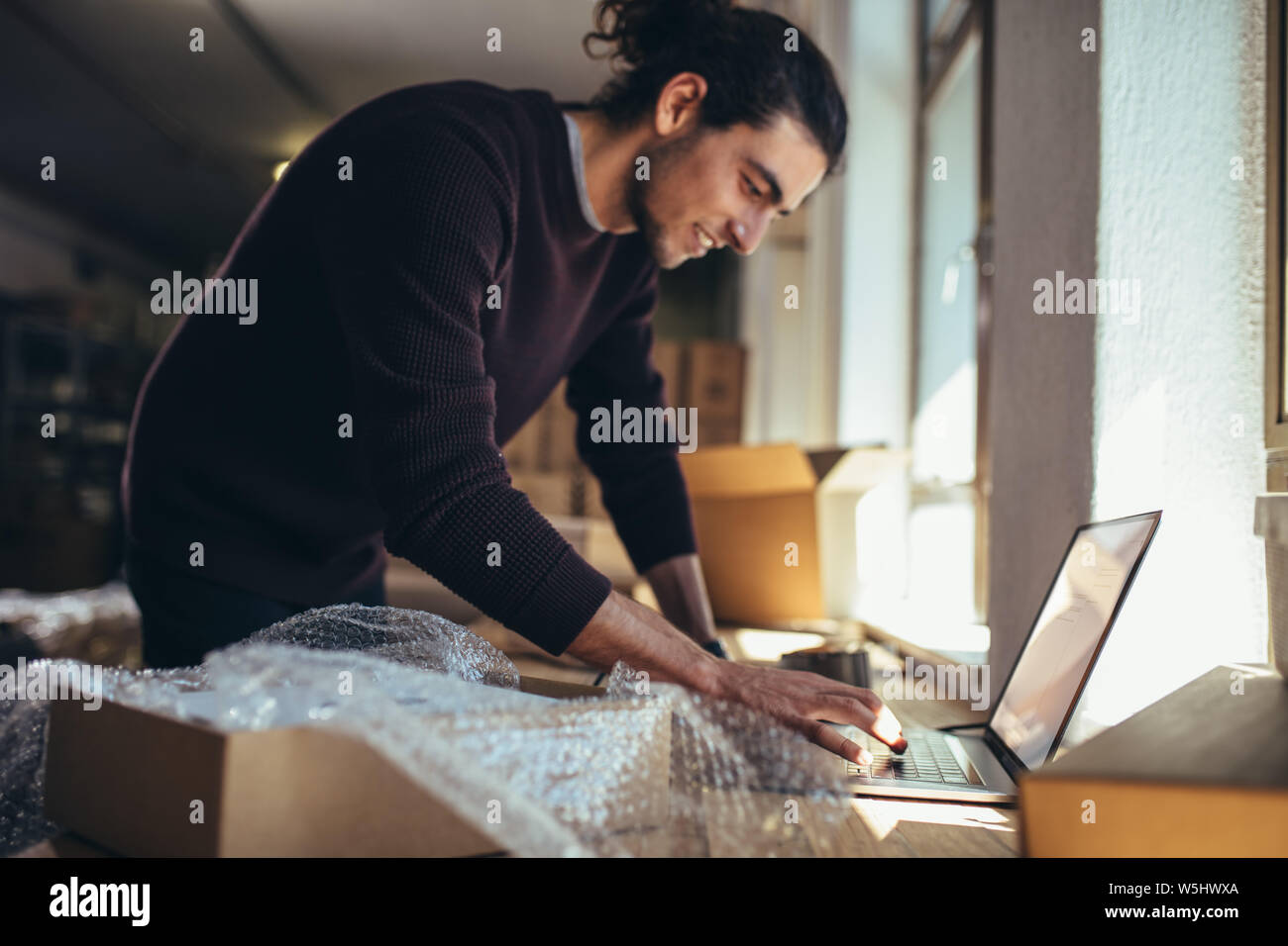 Young man working on laptop with parcel on the side. Drop shipping business owner updating the delivery status of the shipment. Stock Photo