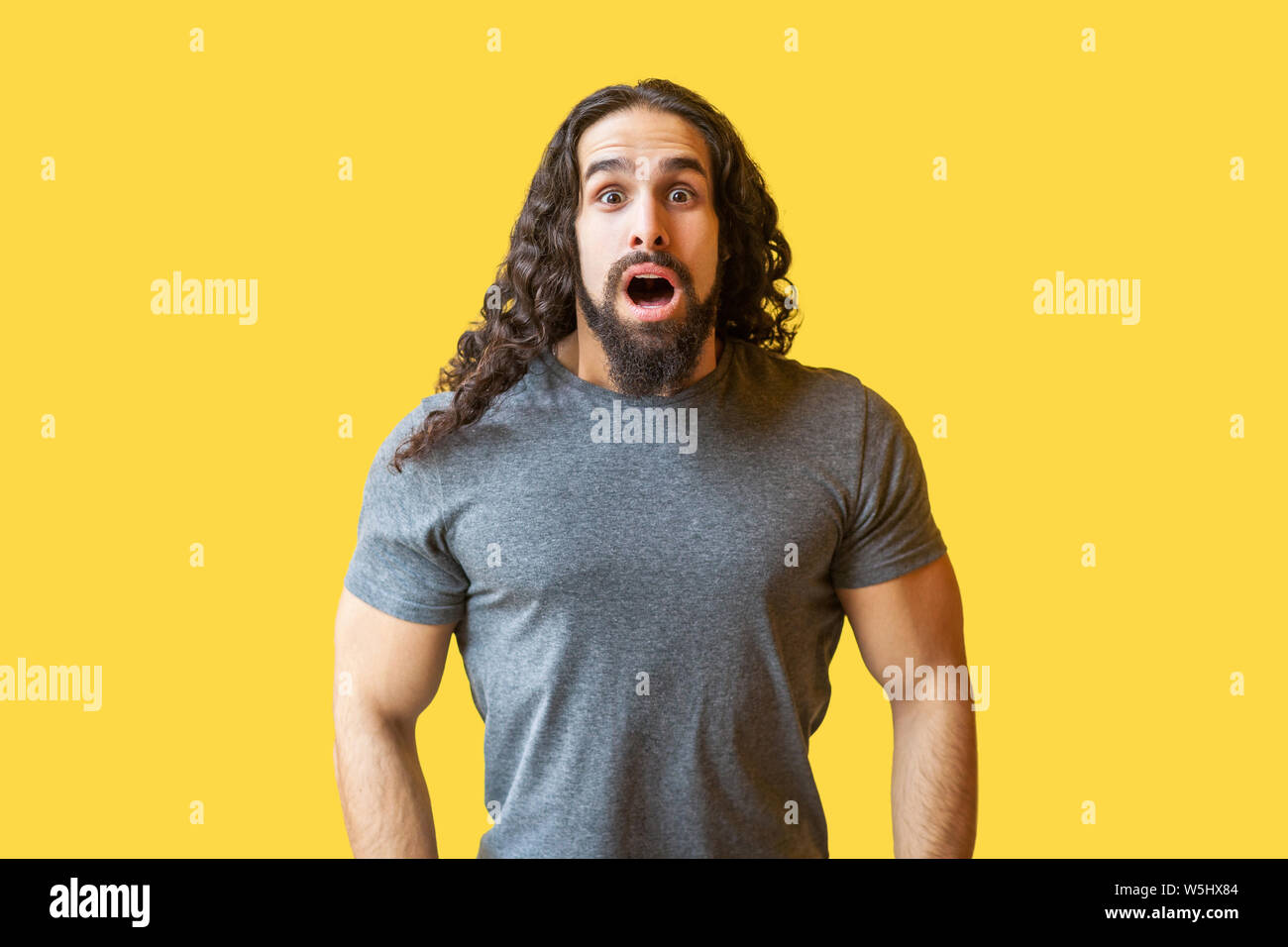 Portrait of shocked bearded young man with long curly hair in grey ...