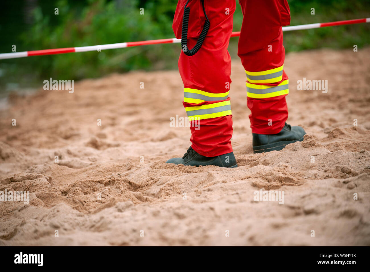 Legs of rescuer standind on the sand in front of red and white tape. Red trousers with reflective elements. Stock Photo