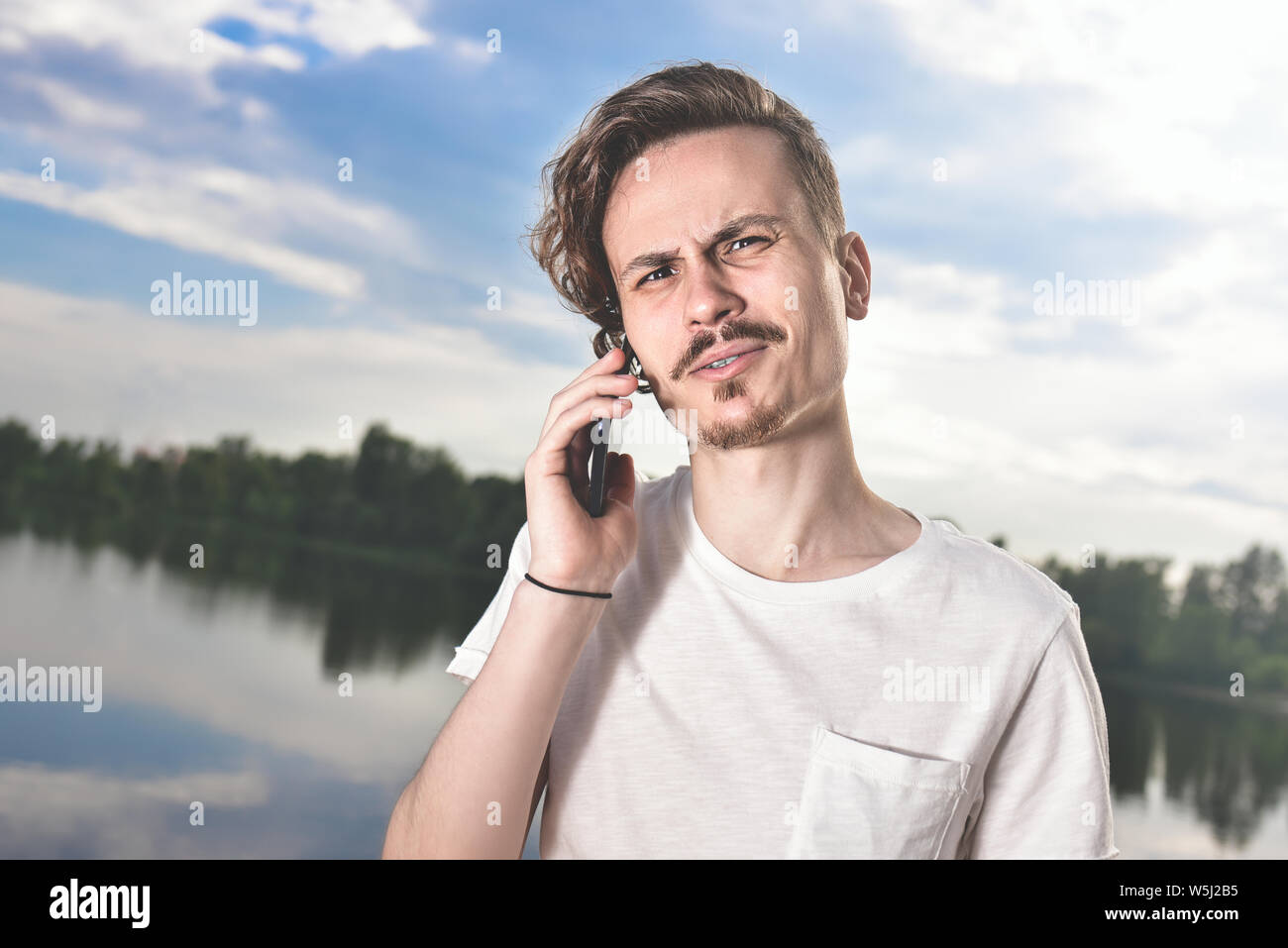 Portrait of angry confused aggressive in bad mood guy irritably speaks on the phone on the background of the lake Stock Photo