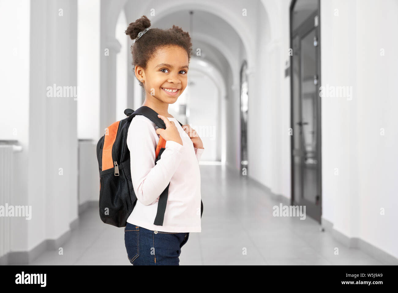 Side view of pretty little African school girl walking in school on break , smiling at camera. Cute and happy girl wearing in white blouse and jeans holding big bag pack. Concept of school. Stock Photo