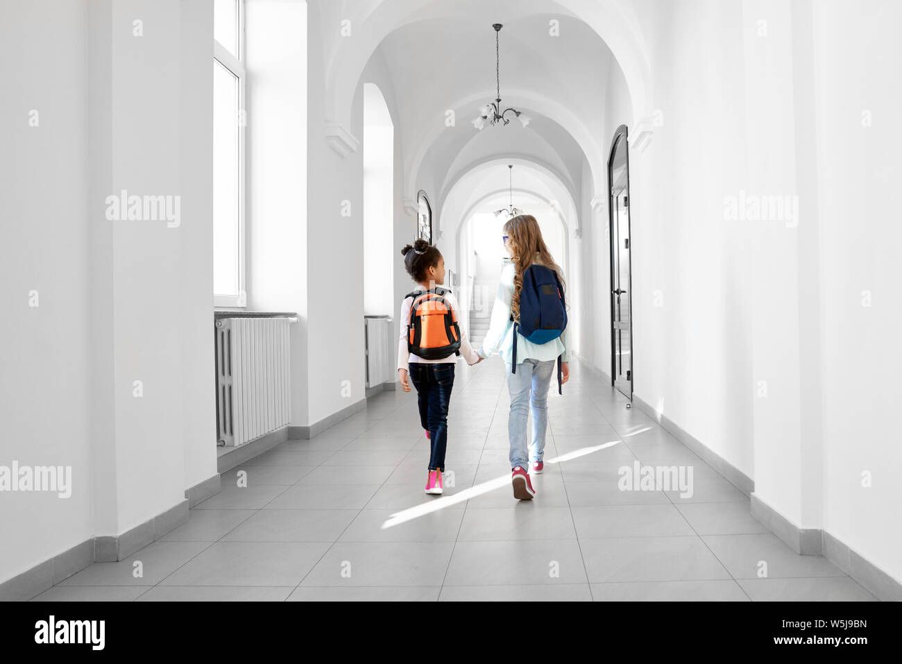 Back view of two girls going forward at school corridor with backpacks on back. Two school girl friends going home from lessons. Concept of study in beautiful school. Stock Photo