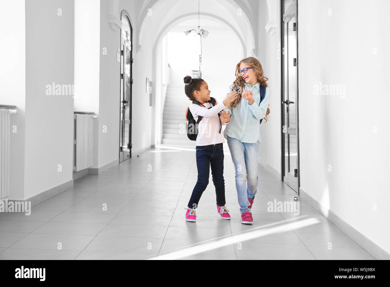 Two funny school girls playing and having fun together, running on long corridor. African and Caucasian girl with different in age studying together in International school. Concept of friendship. Stock Photo