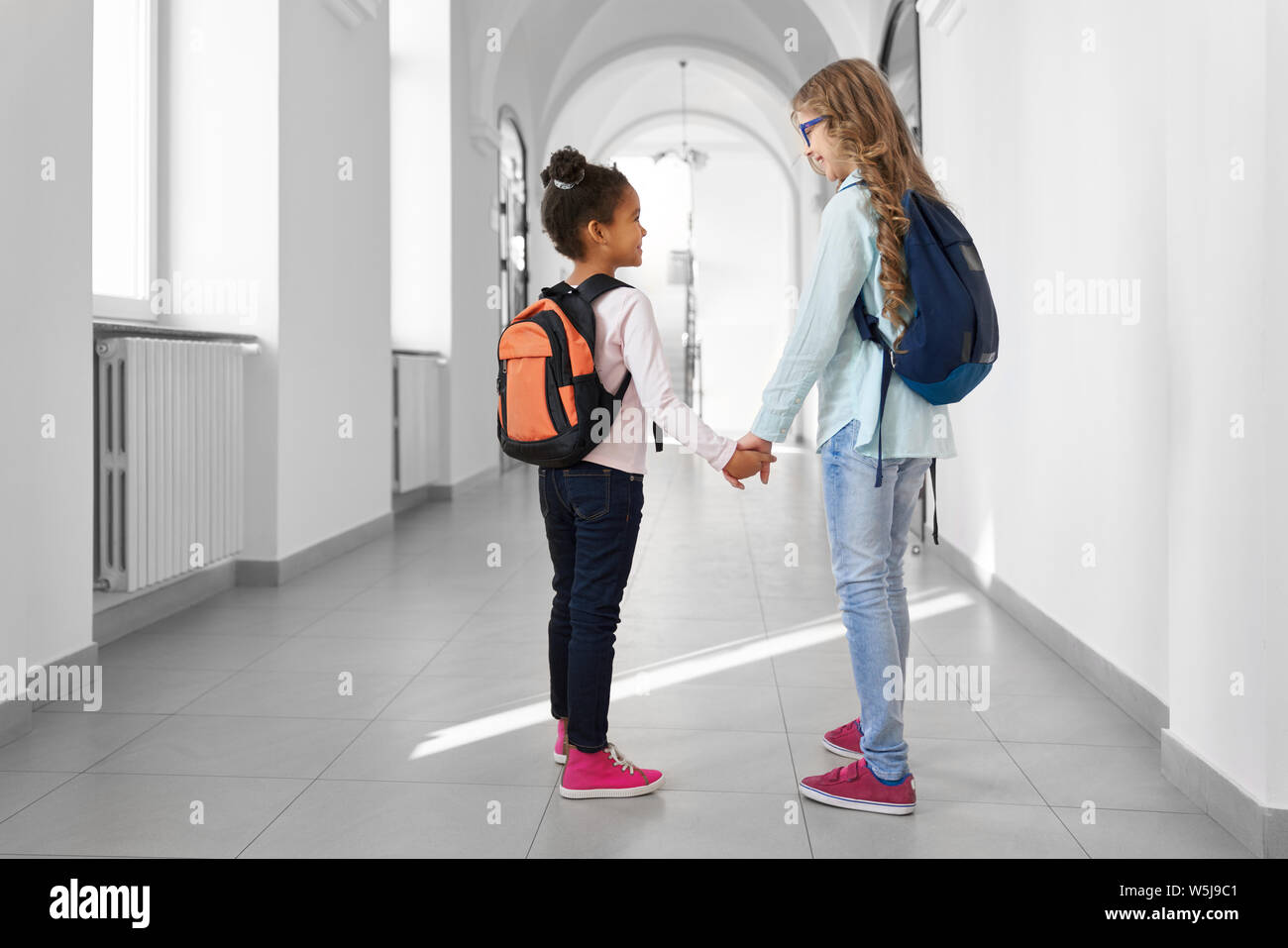 Back view of two school girls African and Caucasian holding by hands going forward at long corridor and looking each other. Two best friends with backpacks walking together at International school. Stock Photo