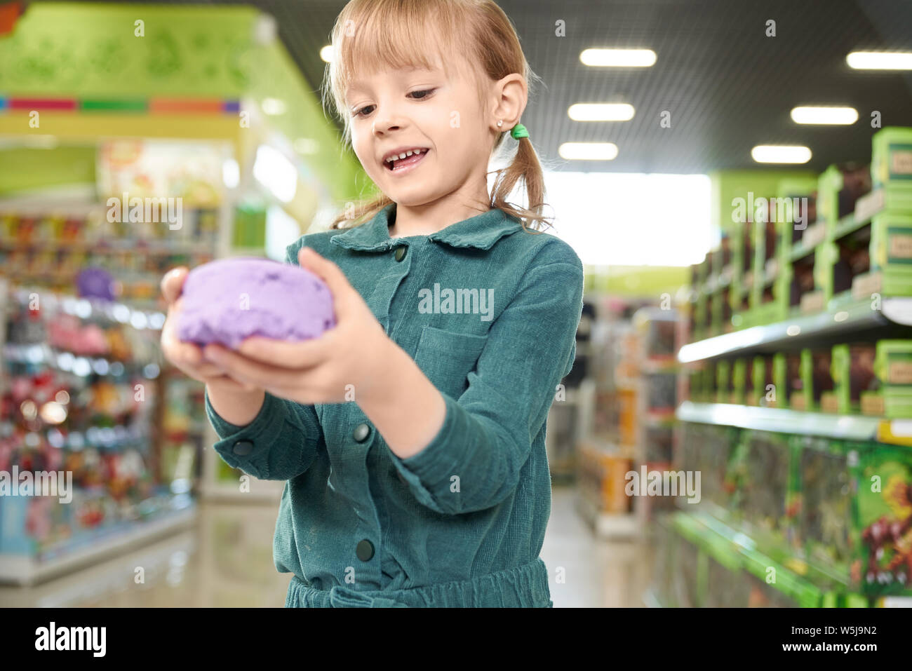 Cute, pretty girl holding in hands violet kinetic sand, posing at camera. Little child playing in playing room in store of shopping centre. Shelves with toys on background. Stock Photo