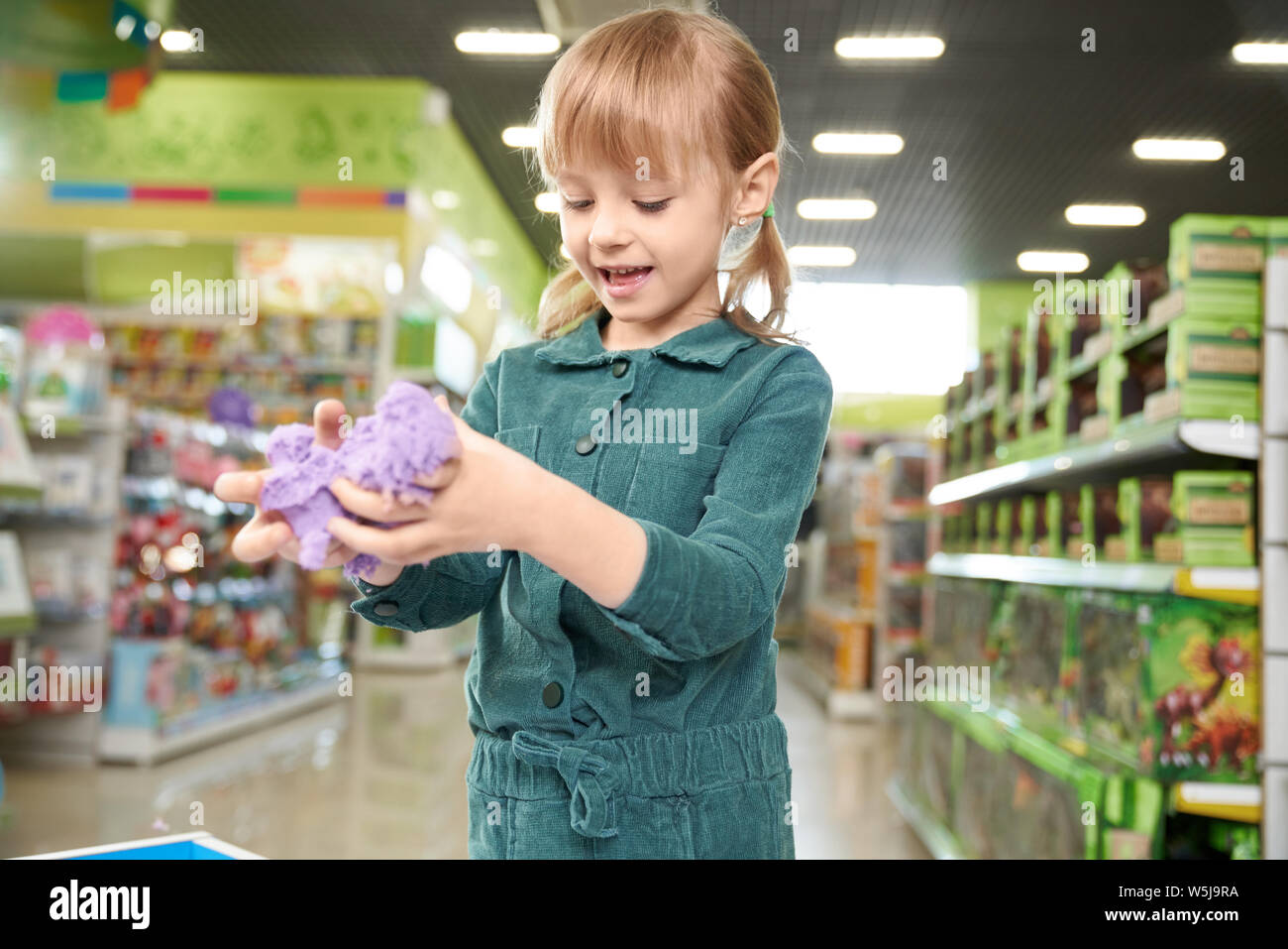 Modern toys for development in store of children toys. Happy pretty girl playing with violet kinetic sand in shopping centre. Child standing in playing room of store. Stock Photo