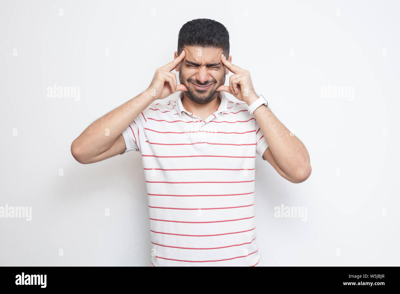 headache or confution. Portrait of confused handsome bearded young man in striped t-shirt standing, holding his painful head or thinking. indoor studi Stock Photo
