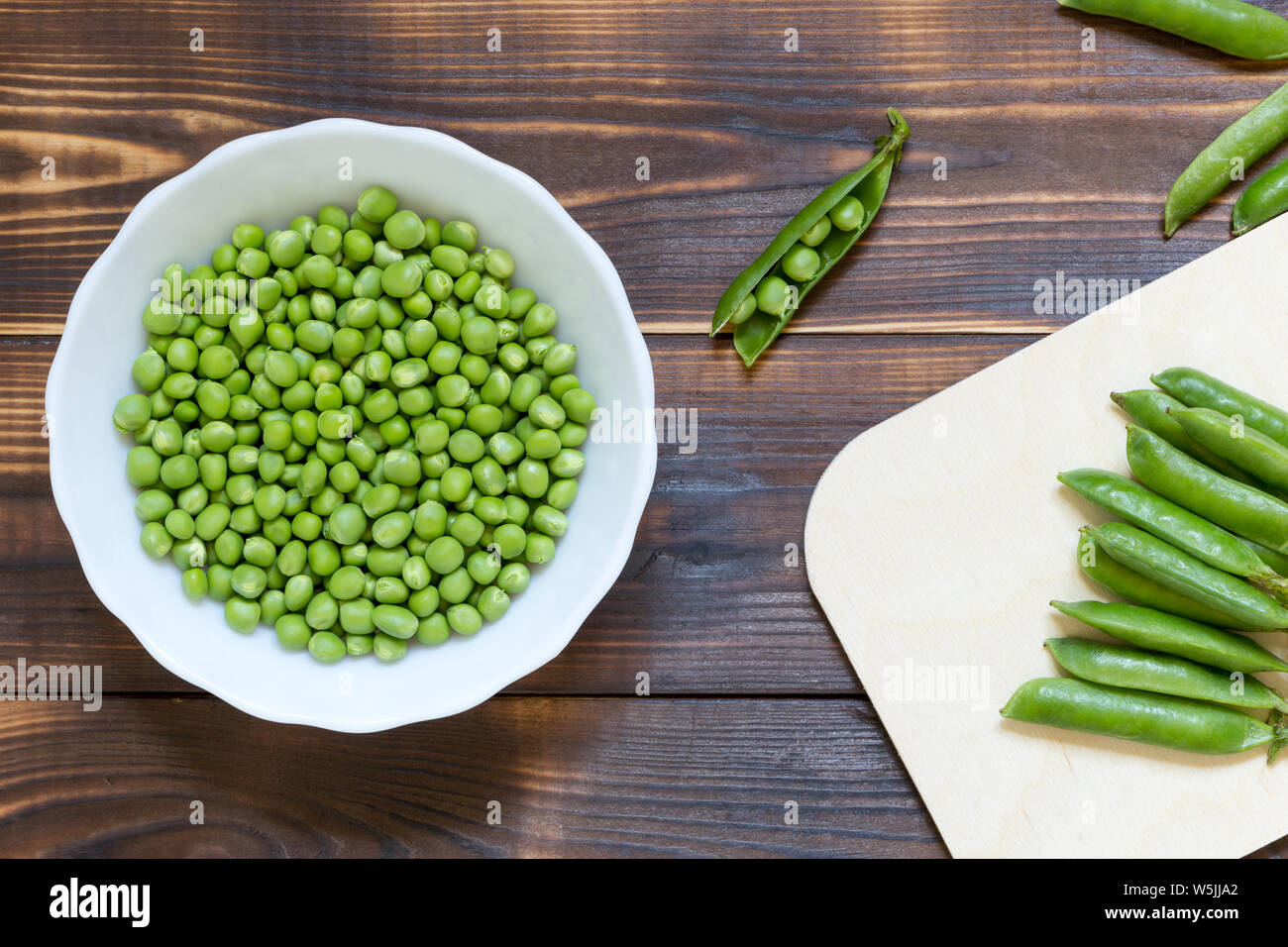 White bowl with green peeled peas inside on dark wooden table. Whole pea pods on kitchen desk and single open pod near. Top view background. Natural o Stock Photo