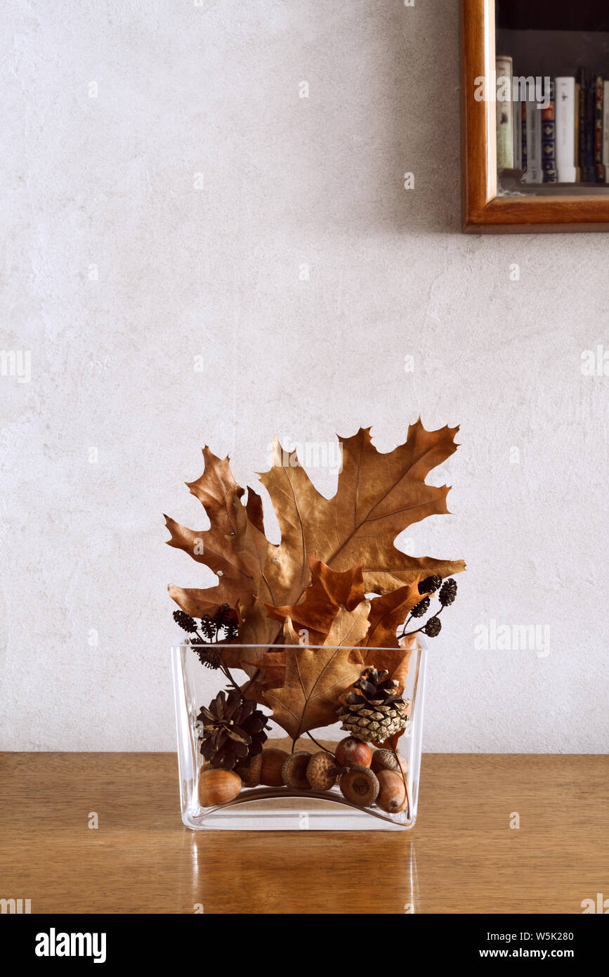 Autumnal home decor of golden and brown leaves, cones and acorns in modern glass vase on table, copy space, partly visible mirror with bookshelf Stock Photo