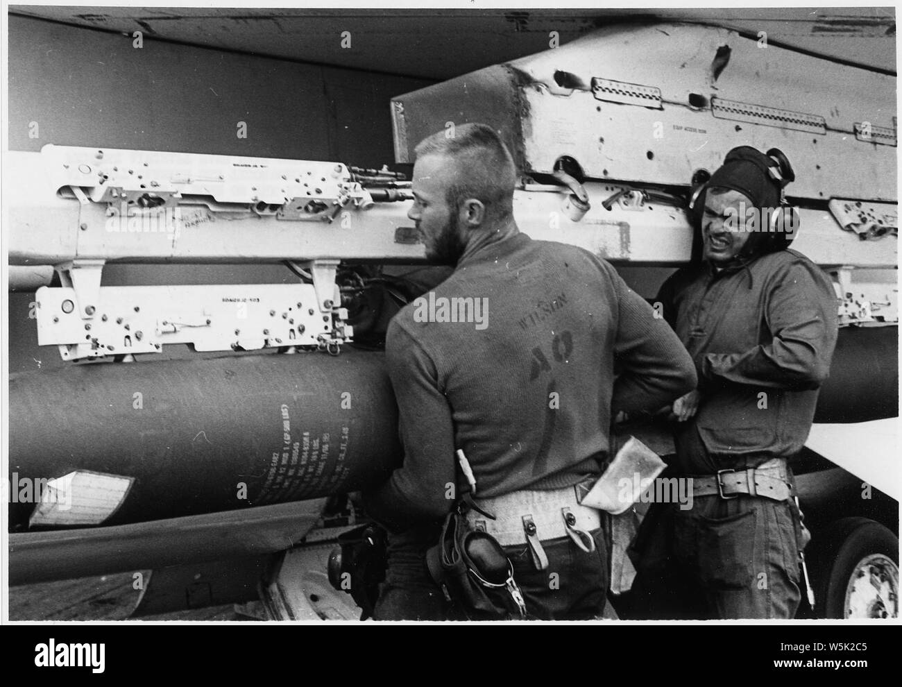 Aviation Ordnanceman Second Class R. P. Wilson of Hinton, West Virginia, (left), and Aviation Ordnanceman Third Class D. A. Bibbee of Portland, Oregon, place a bomb on the rack of an F8 Crusader aboard the attack aircraft carrier USS Ticonderoga (CVA 14). During recent months, the fighter aircraft have been doubling as attack aircraft. Squadron 194 pilots have dropped over 135,000 pounds on enemy targets. Stock Photo