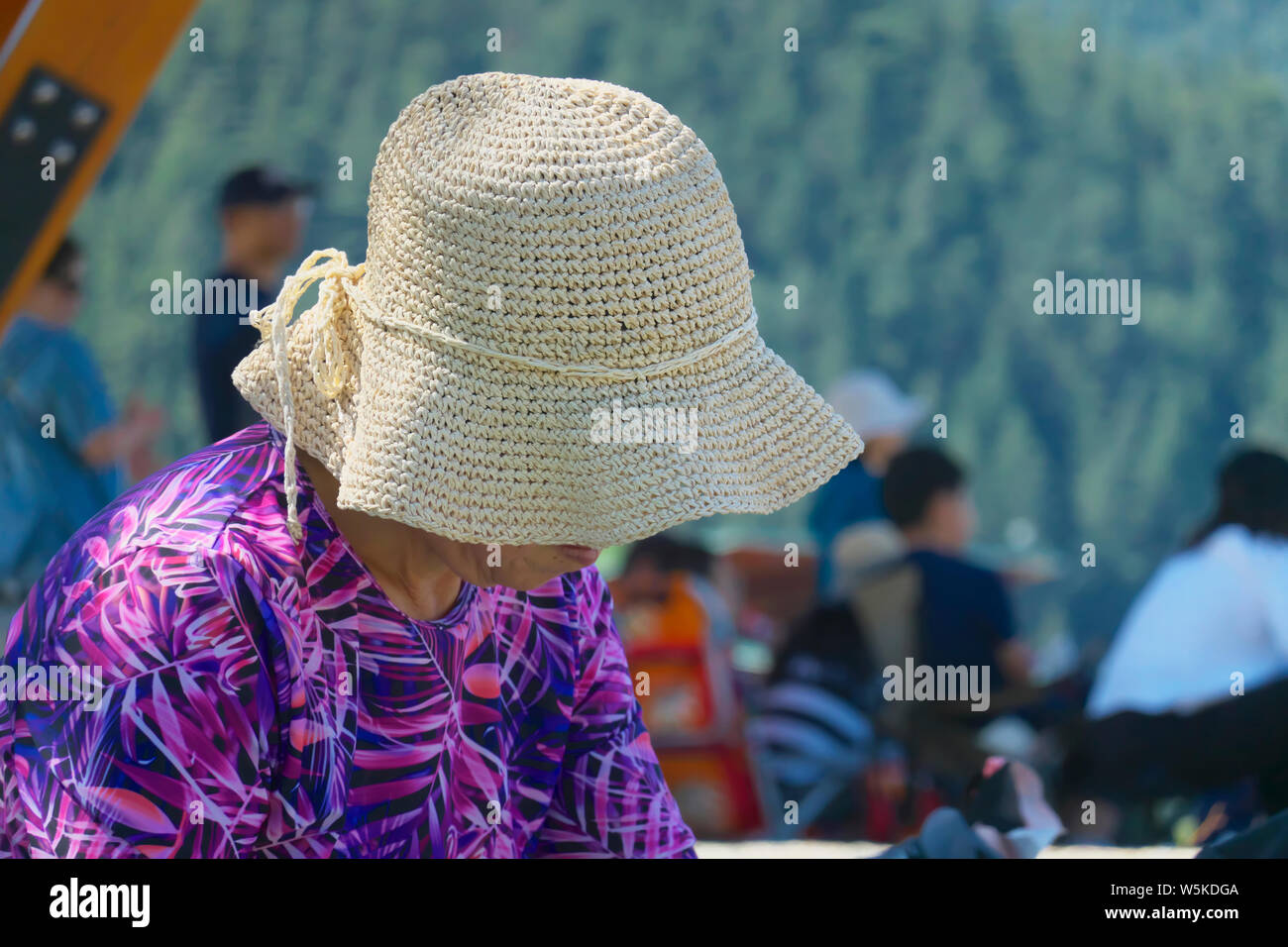 A close-up view of a straw hat on a woman who is wearing a bright purple and pink blouse and looking down. Stock Photo