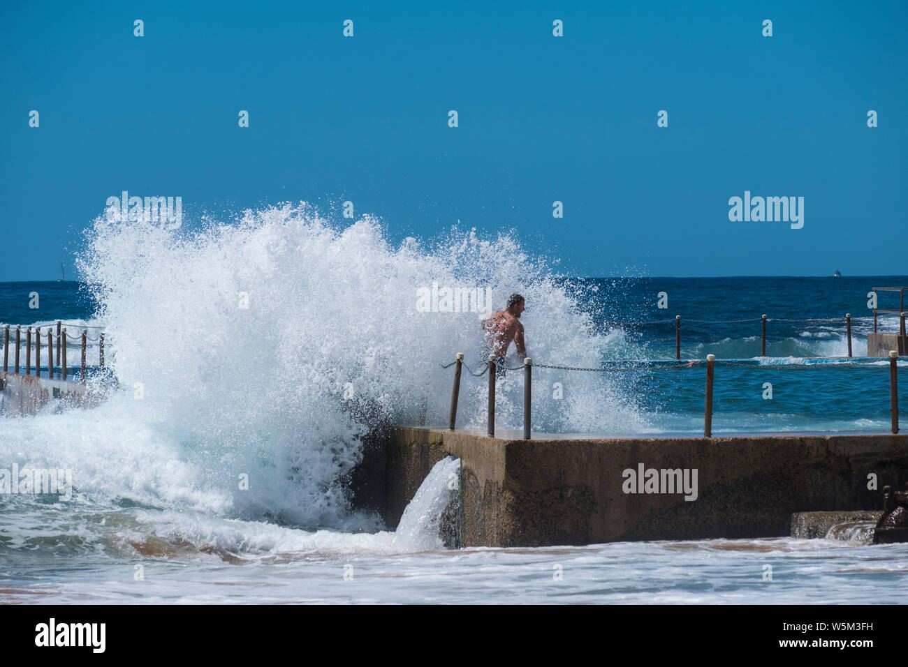 A wave crashes onto a wall of an ocean pool. Bathers enjoys the thrill of the spray. Stock Photo