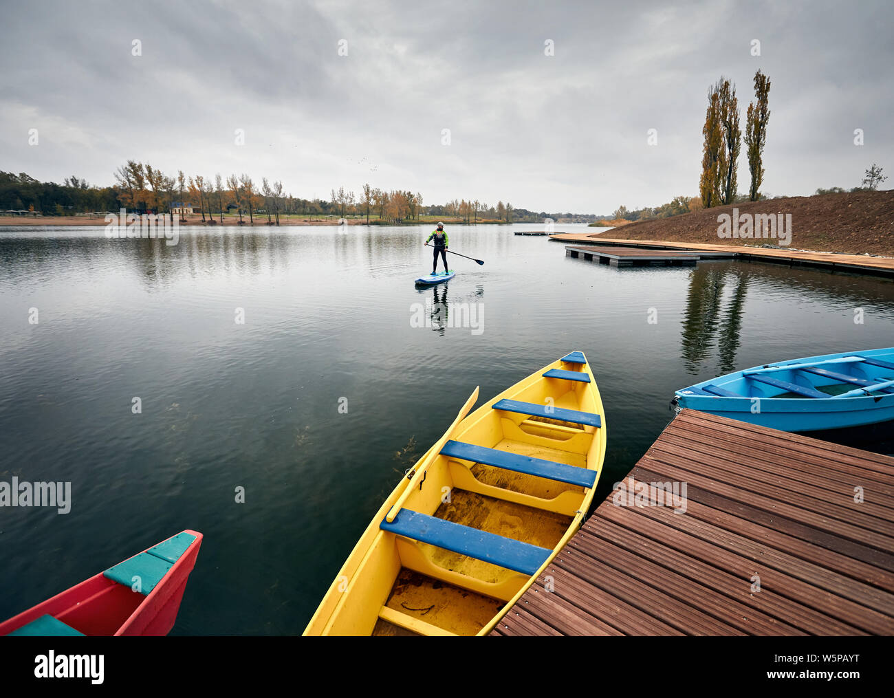 Man on the paddleboard in the lake against overcast sky and pier with colorful boats at foreground Stock Photo