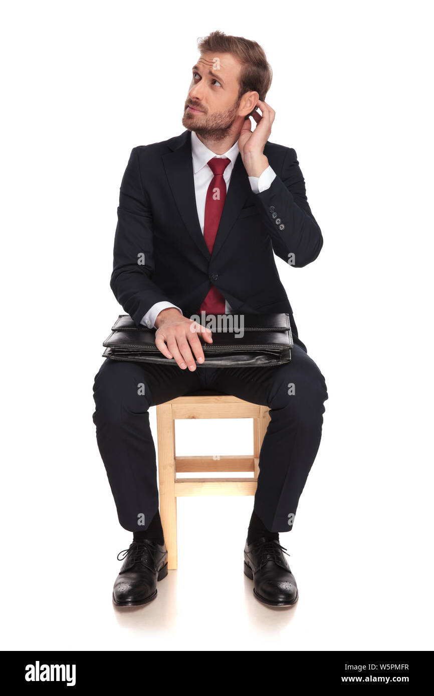 pensive businessman waits for interview and looks up to side while sitting on a wooden chair on white background and holding his head, full length pic Stock Photo