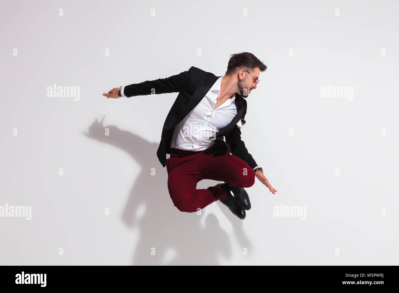 smart casual man jumping and doing a heel click near a white wall while looking down to side, full body picture Stock Photo