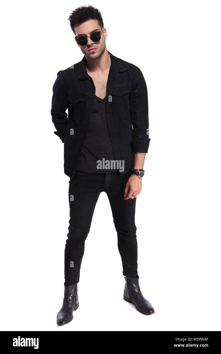 handsome macho man in black clothes and wearing sunglasses standing on white background with a hand behind his back, full length picture Stock Photo