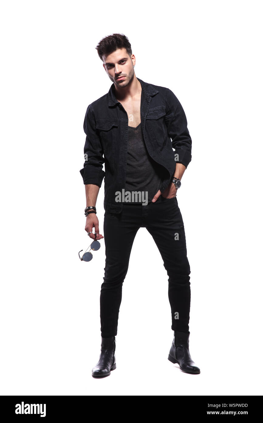 relaxed young man in black clothes standing on white background and holding sunglasses, looking cool and macho, full body picture Stock Photo