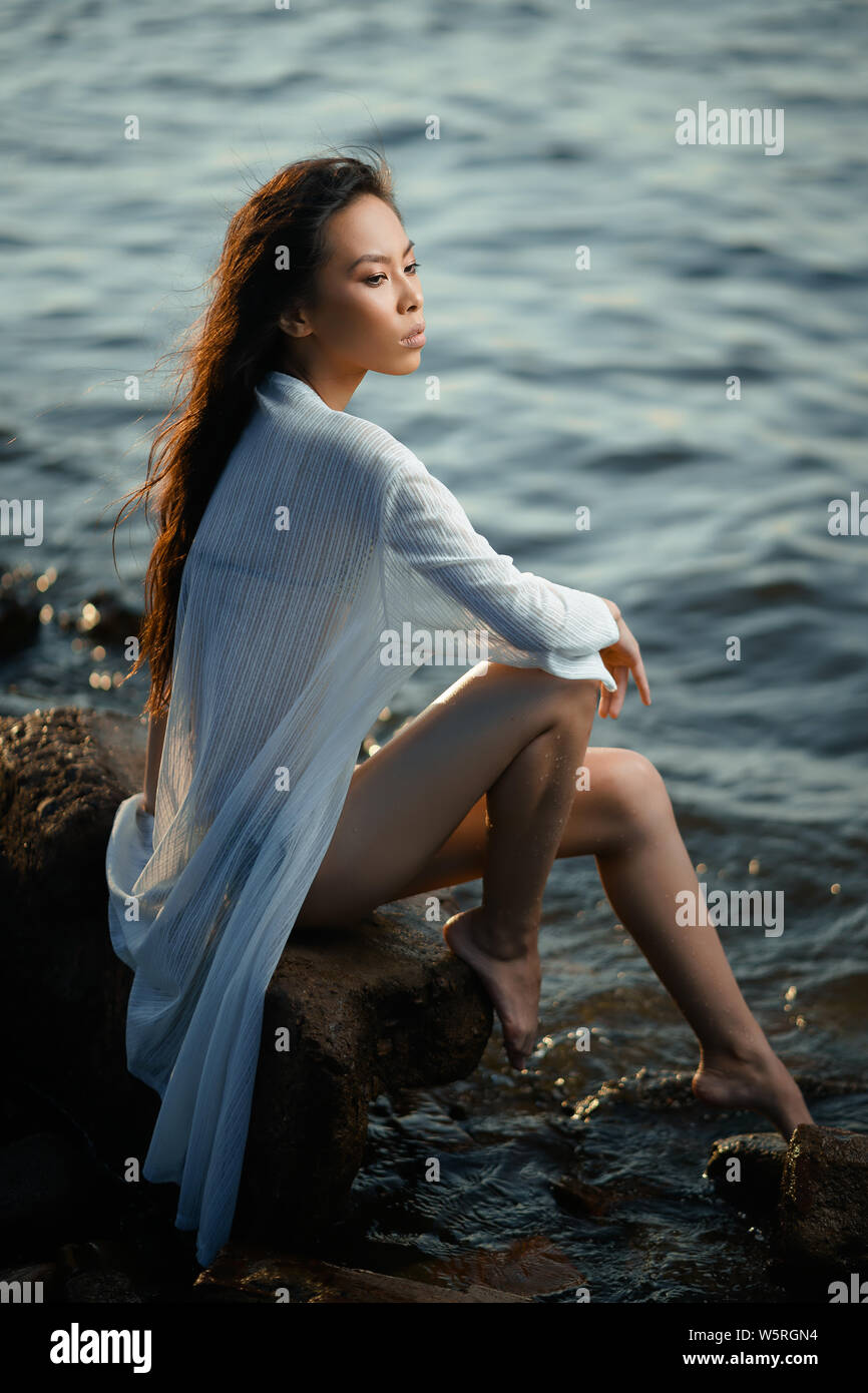 Concept: tourism, holidays, travel. Young lean pretty asian girl in two pieces green swimsuit and long white shirt relax on seashore at sunset. Fashio Stock Photo