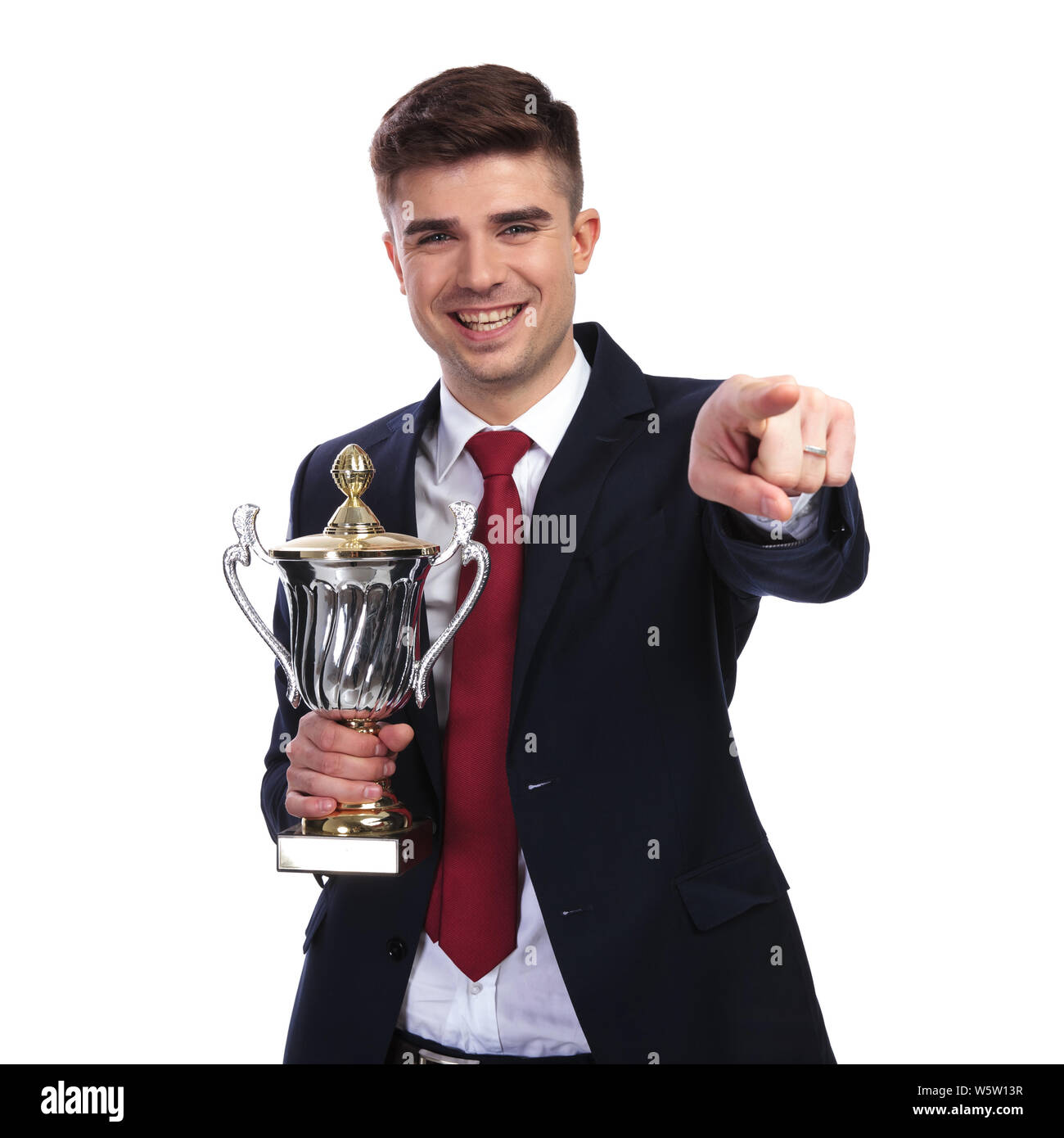 portrait of smiling businessman pointing finger while holding the awarded trophy. He is standing on white background while wearing a red tie and a nav Stock Photo