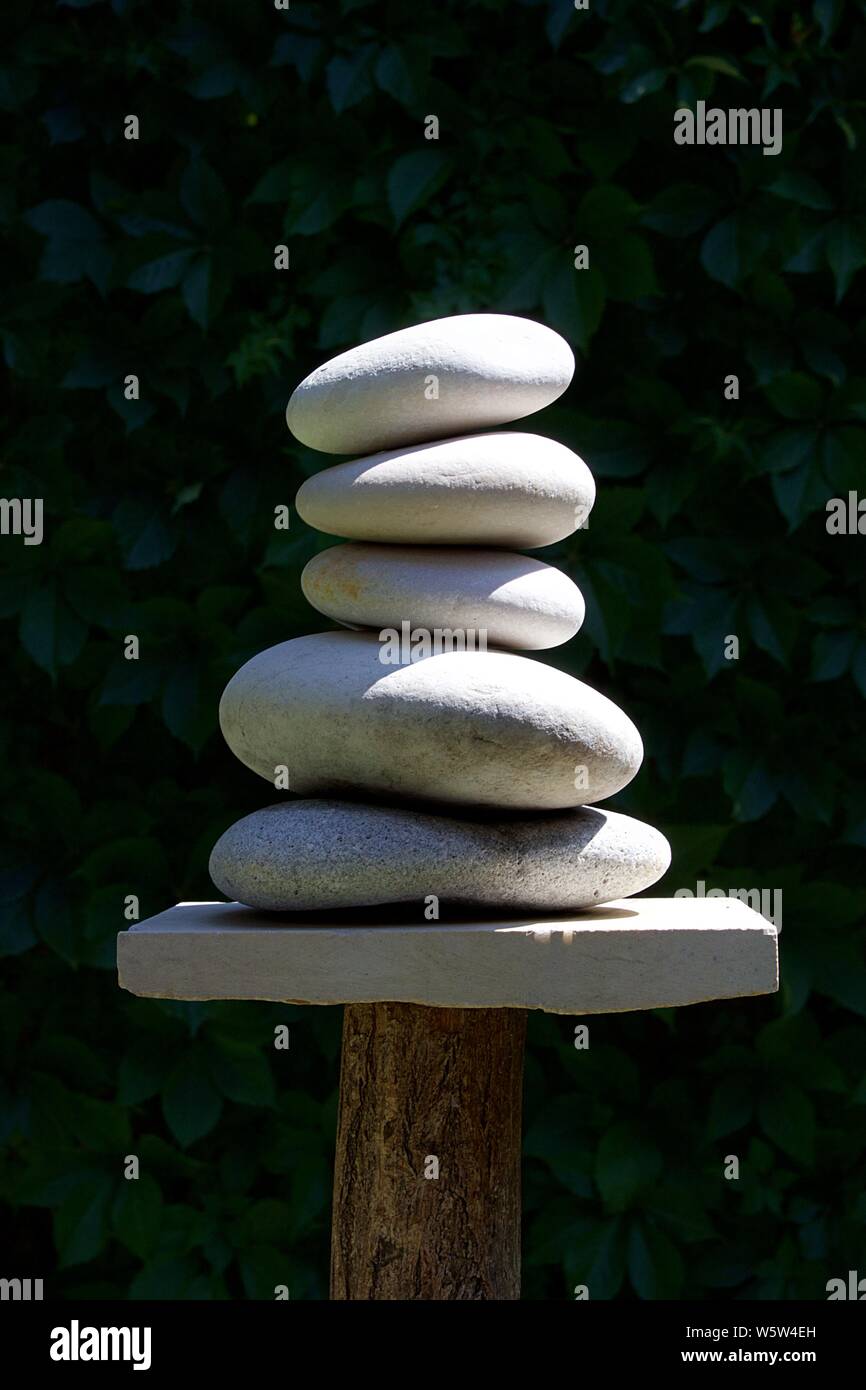 Zen sculpture of five white pebbles cairn tower a marble and a log. Blurred background of dark green foliage Stock Photo