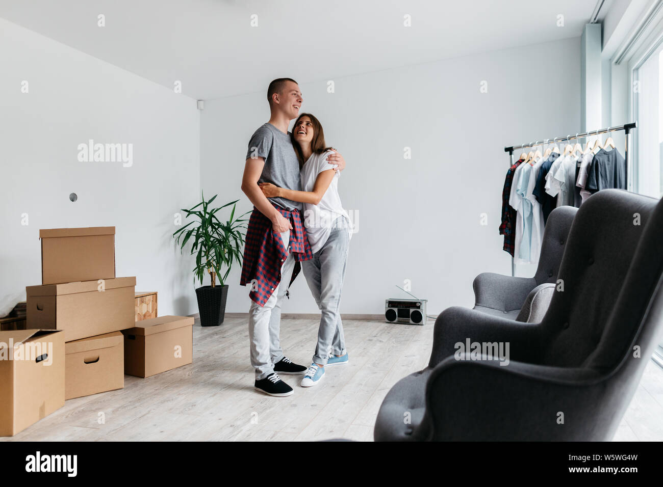Happy couple hugging together in new house. Moving in together - young cheerful man and woman cuddling in room with cardboard boxes. Stock Photo