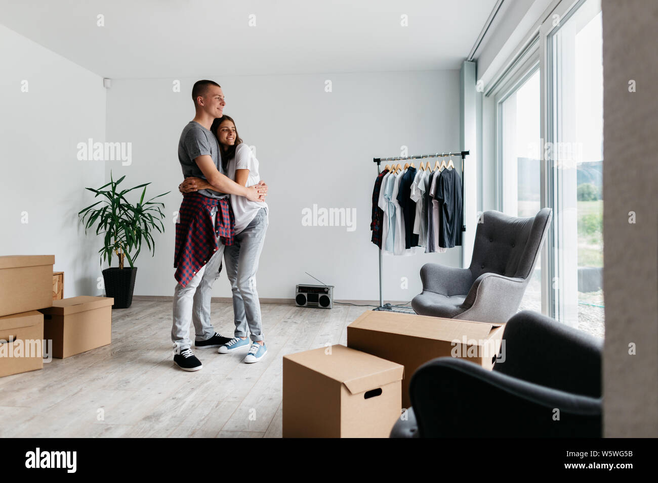 Happy couple hugging together in new house. Moving in together - young cheerful man and woman cuddling in room with cardboard boxes. Stock Photo