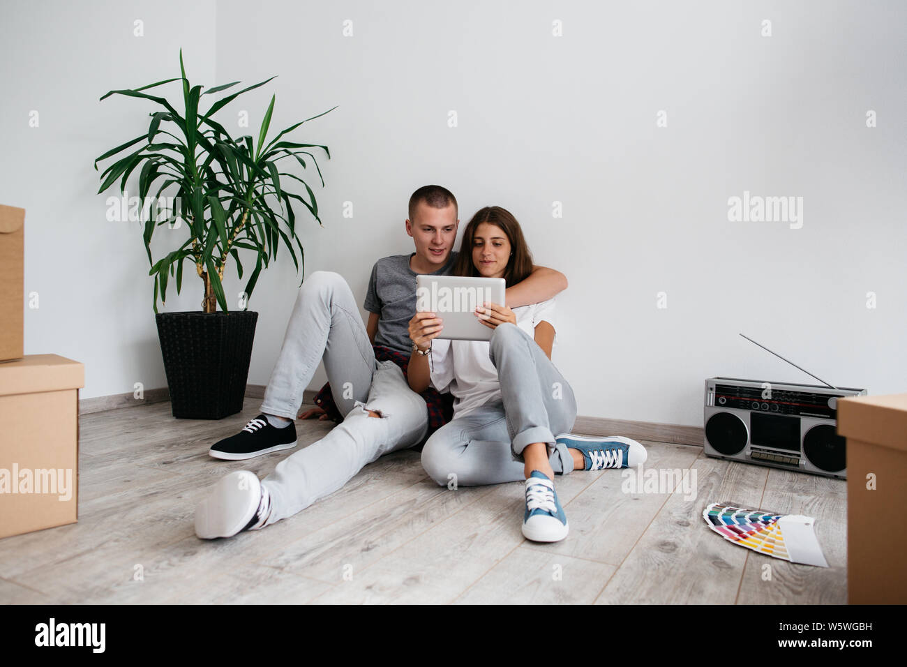 Moving in - couple in love sitting on floor in empty room of their new house. Young man and woman planning design of their new home with digital table Stock Photo