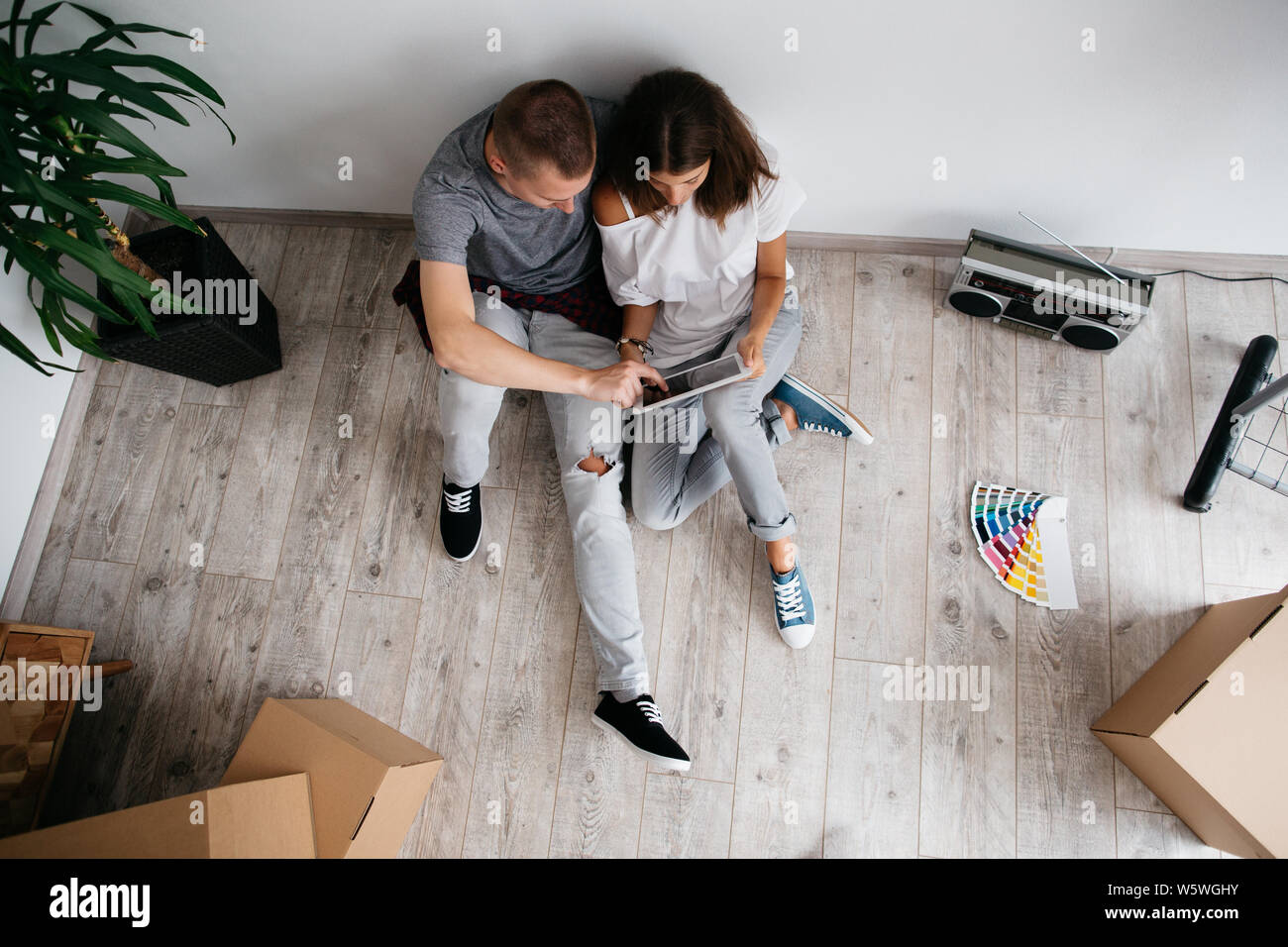 Relocation - couple sitting on floor in empty room of their new house. Top view of young man and woman planning design of their new home with digital Stock Photo
