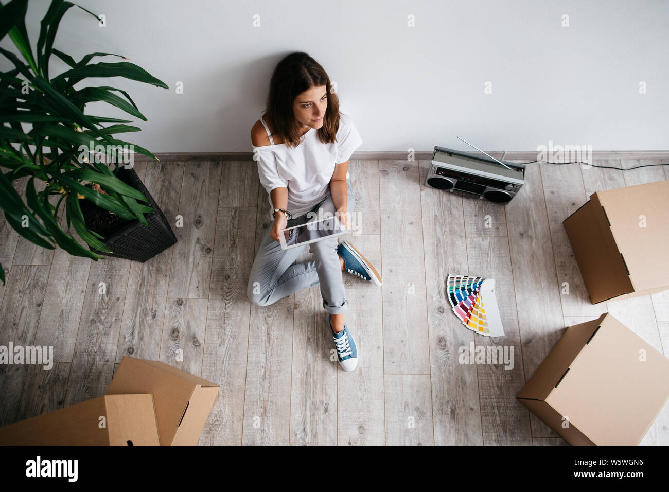 Young woman sitting on floor in empty room of her new house. High angle view of woman planning interior of her new home with digital tablet and color Stock Photo