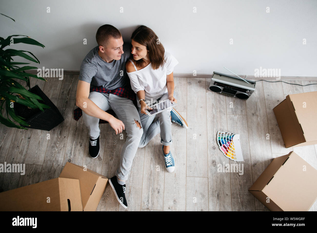 Couple in love sitting on floor in empty room of their new house. High angle view of young man and woman planning interior of their new home with digi Stock Photo