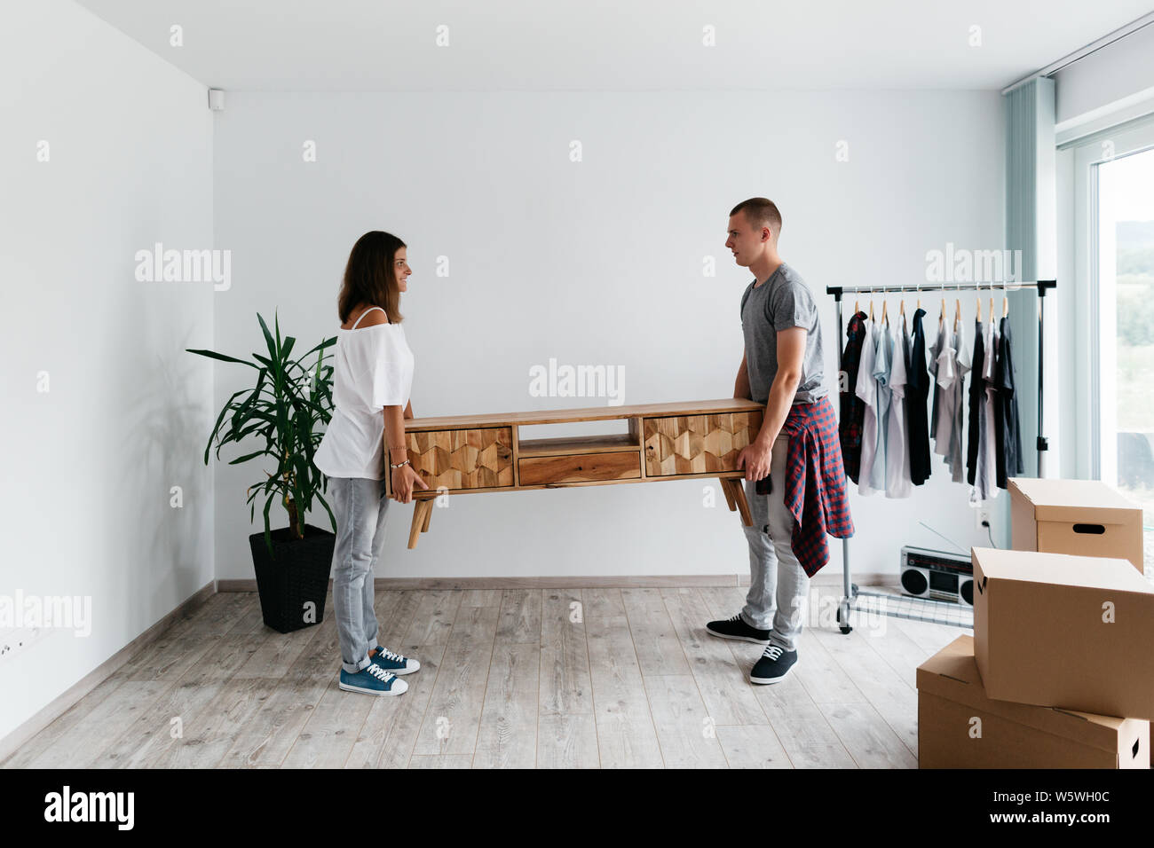 Couple moving furniture in their new house. Relocation - young man and woman looking at each other whilst carrying wooden TV cabinet against white wal Stock Photo
