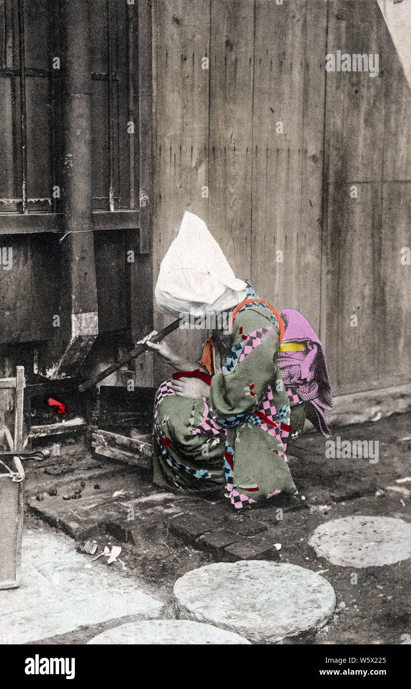 [ 1900s Japan - Japanese Woman Heating Bathwater ] —   A young woman in kimono is tending a fire to heat bathwater with a hifukidake (火吹き竹), a bamboo tube used to kindle a fire.  20th century vintage postcard. Stock Photo