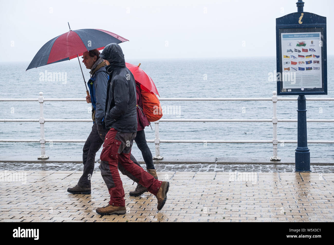 Aberystwyth, Wales, UK. 30th July 2019.  Holidaymakers  in their raincoats and umbrellas walking along the promenade in Aberystwyth in the rain as a belt of thundery and very wet weather spreads across much of the central parts of the UK, bringing with it the risk of disruptive flooding and treacherous driving conditions.. Photo credit : Keith Morris/Alamy Live News Stock Photo