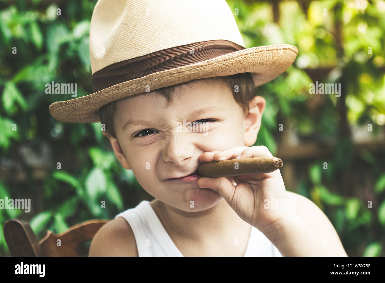 Beautiful young boy plays imitating an adult who smokes a cigar. Funny kid makes faces by wrinkling his nose while holding a cigar in his hand. Close Stock Photo