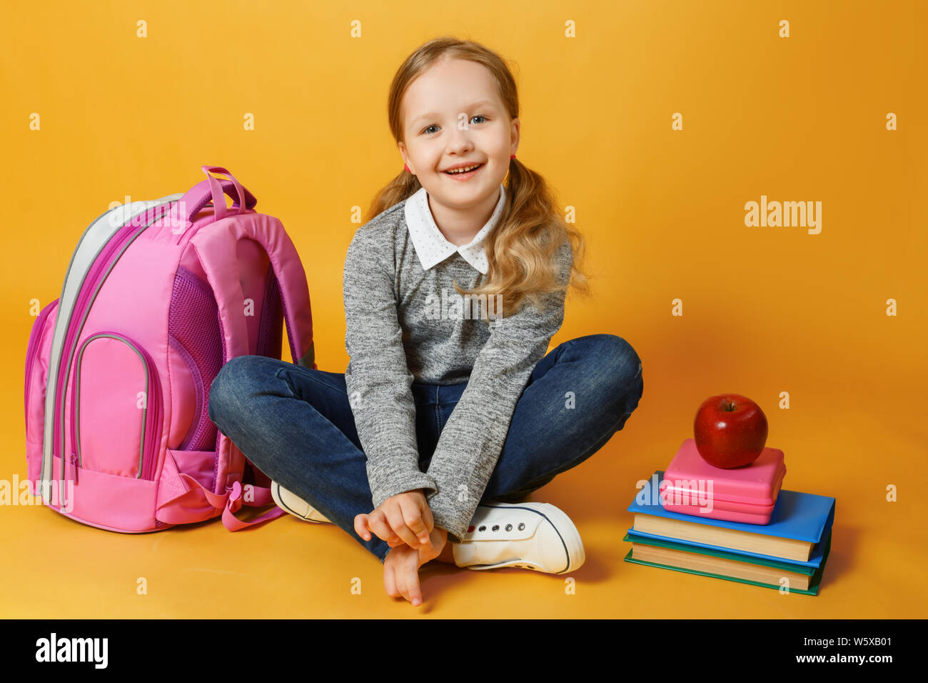 Cheerful little student girl is sitting with backpack, books and lunch box on yellow background. Child student looks into the camera. The concept of e Stock Photo