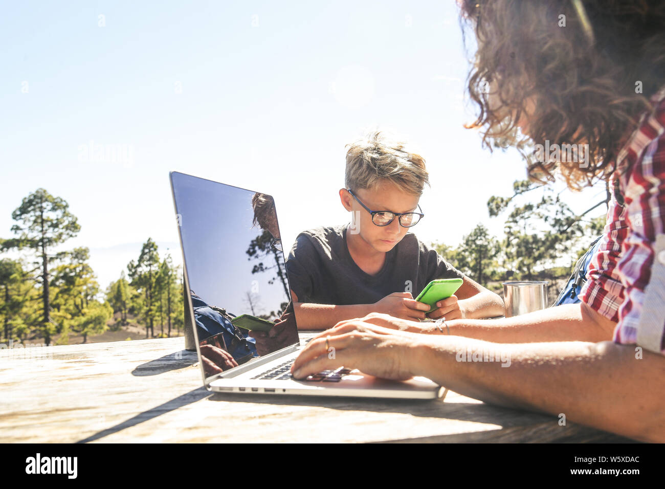 Close up view of mother and son taking rest after mountain trekking Mum working with laptop Trendy young boy use smartphone Blogger girl making social Stock Photo