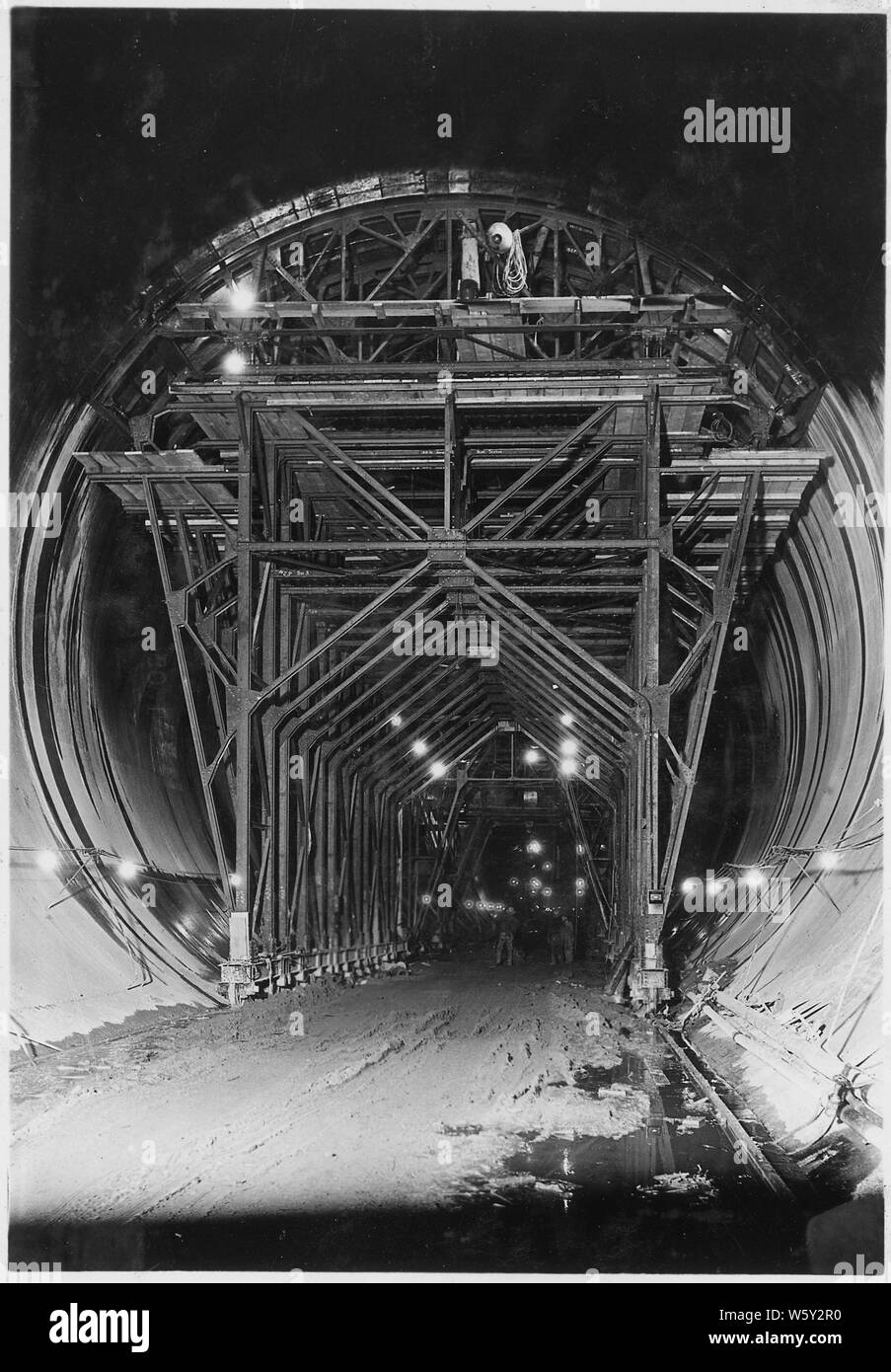 Steel frame carriage supporting form for 110 degree top-arch concrete in diversion tunnel lining. Note jacks and wedges by means of which form is placed in position and lined up. The surface of the top concrete is painted with an asphaltic coating immediately upon removal of the form to prevent the too rapid dissipation of the concrete's moisture content. See also No. 1303 for further description.; Scope and content:  Photograph from Volume Two of a series of photo albums documenting the construction of Hoover Dam, Boulder City, Nevada. Stock Photo