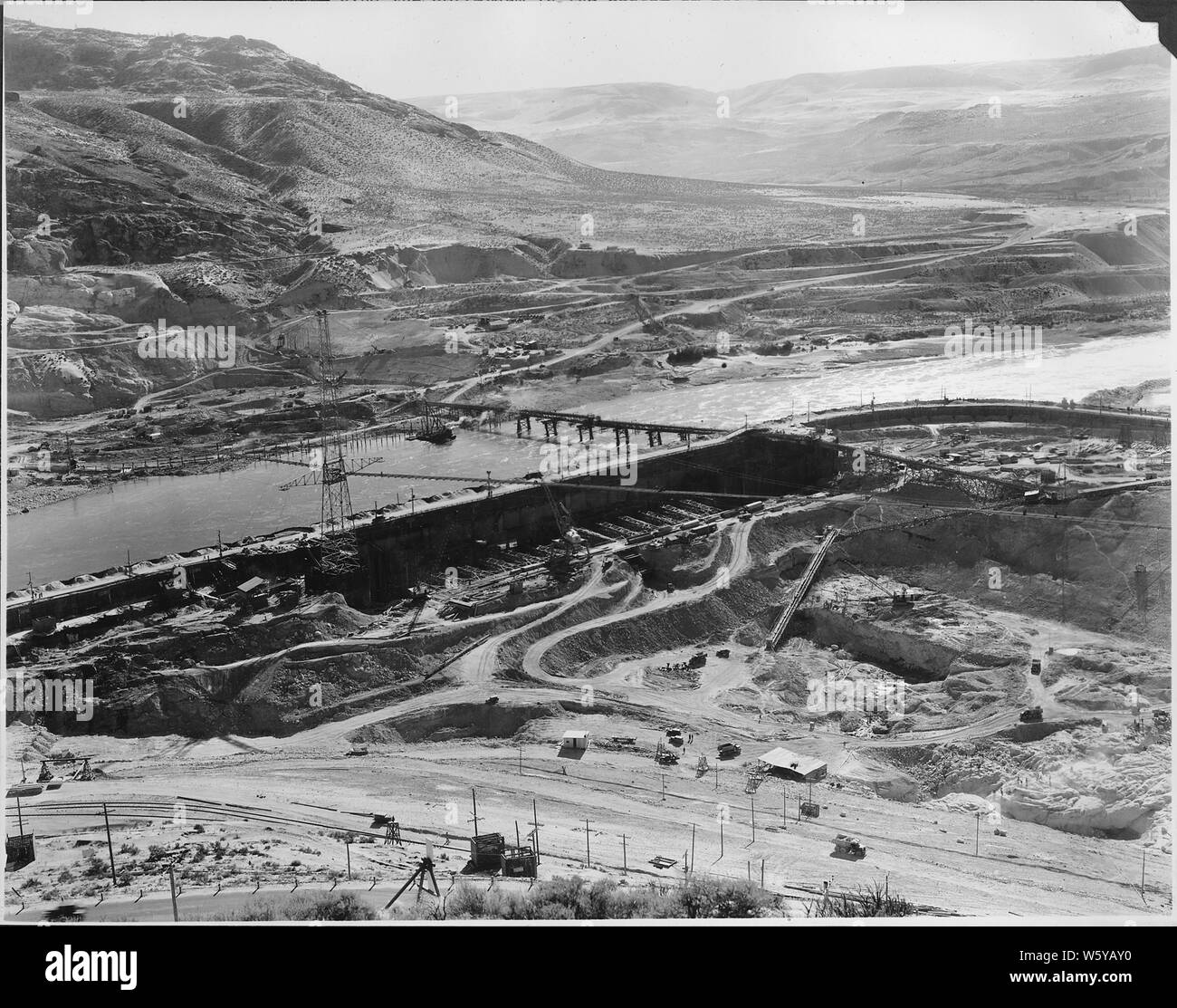 The southeast corner of the immense 3,000 foot Grand Coulee cofferdam; Scope and content:  Photograph from Volume Two of a series of photo albums documenting the construction of the Grand Coulee Dam and related work on the Columbia Basin Project.  Full caption for this photograph reads: The southeast corner of the immense 3,000 foot Grand Coulee cofferdam; between the cell clusters will be seen the timber struts in the section now being excavated for placing the first concrete in the Grand Coulee Dam; overhead is a portion of the 3,500 foot suspension bridge for transporting sand and gravel fr Stock Photo