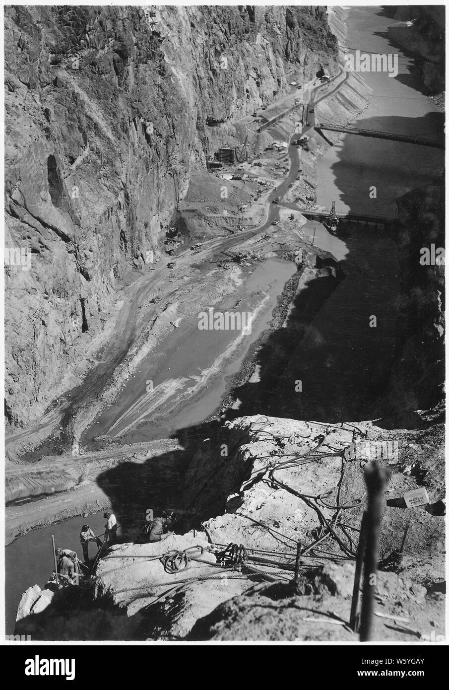 View looking into Black Canyon from rim of the Arizona wall showing operations in the early stages of the upper cofferdam construction; Scope and content:  Photograph from Volume Two of a series of photo albums documenting the construction of Hoover Dam, Boulder City, Nevada.  Full caption of photograph reads: View looking into Black Canyon from rim of the Arizona wall showing operations in the early stages of the upper cofferdam construction. Temporary cofferdam diverts river flow through Arizona side of channel. Nevada side inside temporary cofferdam is partially unwatered and muck is being Stock Photo