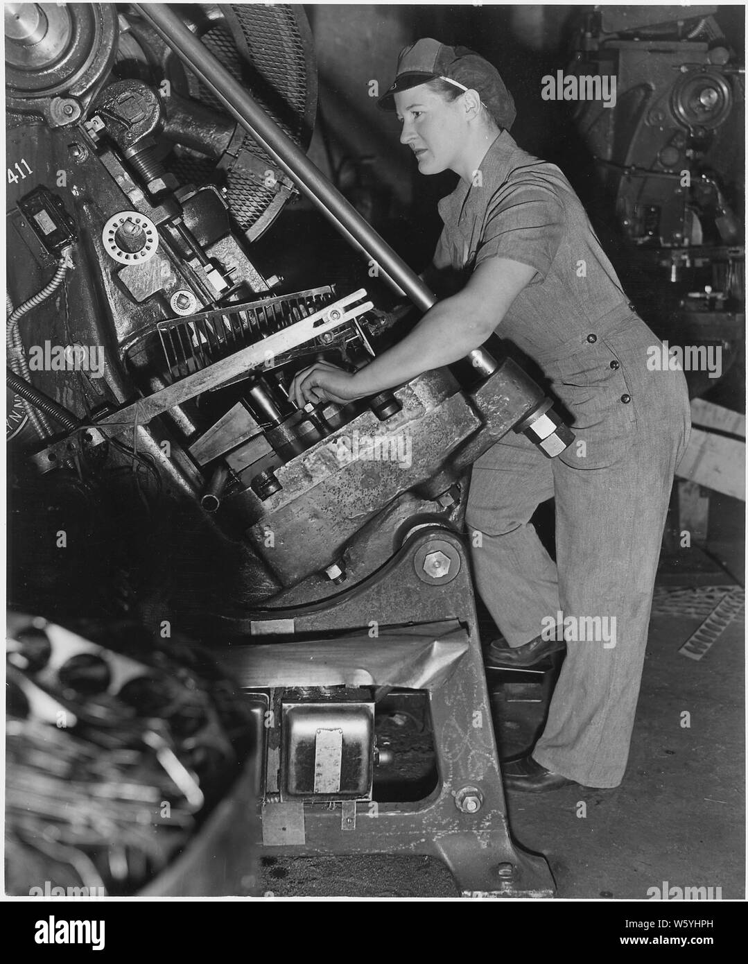 Virginia Ludwig goes to work on a punch press, stamping out discs from steel strips. Her well-fitting uniform is devoid of unecessary pleats, trimmings, doodads - is just right for the job. Stock Photo