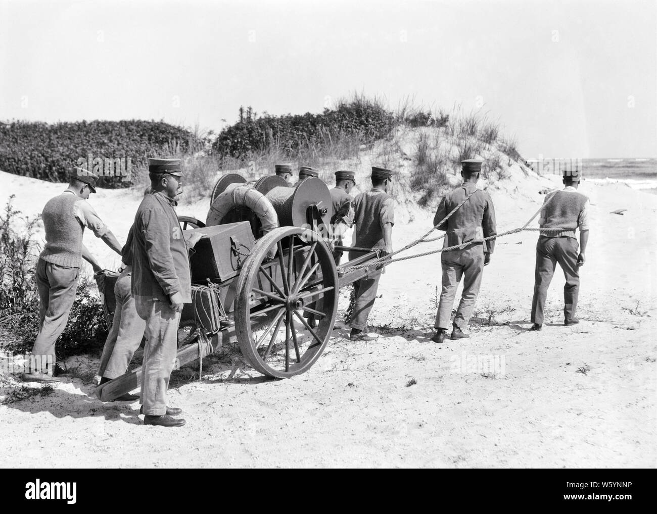 1910s 1920s UNIFORMED COAST GUARD MEN TOWING LYLE GUN AND BREECHES BUOY LIFE SAVING EQUIPMENT ON CART OVER BEACH SAND NJ USA - c98 HAR001 HARS RISK CONFIDENCE TRANSPORTATION B&W LAW ENFORCEMENT NORTH AMERICA NORTH AMERICAN DISASTER ADVENTURE PROTECTION STRENGTH COURAGE AND DUNES EXCITEMENT INNOVATION PRIDE IN ON AUTHORITY NJ OCCUPATIONS SAILORS USING CONNECTION TOWING CONCEPTUAL SQUAD 1790 1848 ESCAPE LONG BEACH ISLAND STYLISH SUPPORT NEW JERSEY SHIPWRECKED UNIFORMED GOVERNMENT AGENCY MARINERS COOPERATION DEPLOYED FORMED LIFESAVING MID-ADULT MID-ADULT MAN MISSION SITUATIONS SOLUTIONS Stock Photo