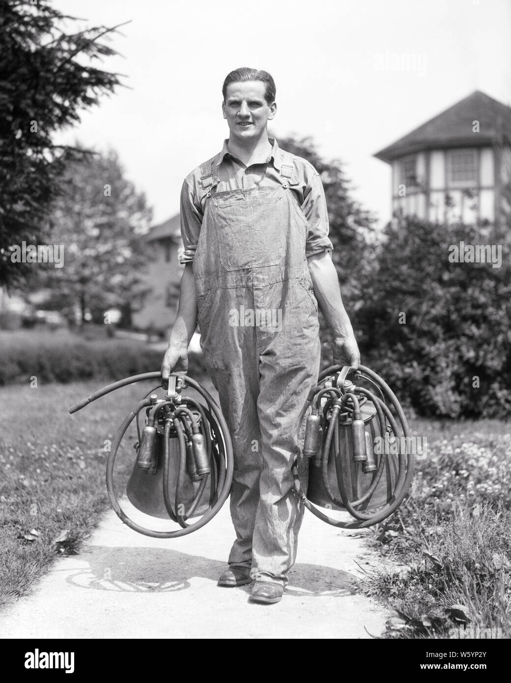 1920s 1930s SMILING MAN DAIRY FARMER WEARING OVERALLS WALKING TOWARD LOOKING AT CAMERA CARRYING TWO VACUUM MILKING MACHINES - d2516 HAR001 HARS DAIRY COPY SPACE FULL-LENGTH PERSONS OVERALLS FARMING MALES PROFESSION AGRICULTURE B&W EYE CONTACT SKILL OCCUPATION SKILLS CAREERS FARMERS PROGRESS TOWARD INNOVATION OCCUPATIONS CONCEPTUAL BAREHEADED MID-ADULT MID-ADULT MAN BLACK AND WHITE CAUCASIAN ETHNICITY HAR001 OLD FASHIONED Stock Photo