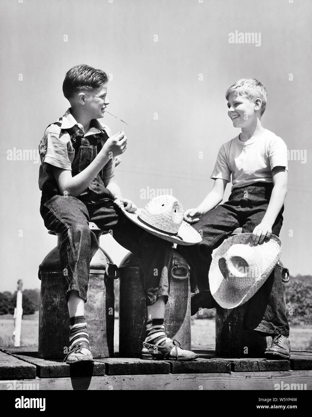 1940s TWO FARM BOYS SITTING ON MILK CHURNS TALKING SMILING WEARING DENIM  HOLDING STRAW HATS BRUNETTE BOY WEARING BIB OVERALLS - d1178 HAR001 HARS  DAIRY COPY SPACE FRIENDSHIP HALF-LENGTH OVERALLS CANS FARMING