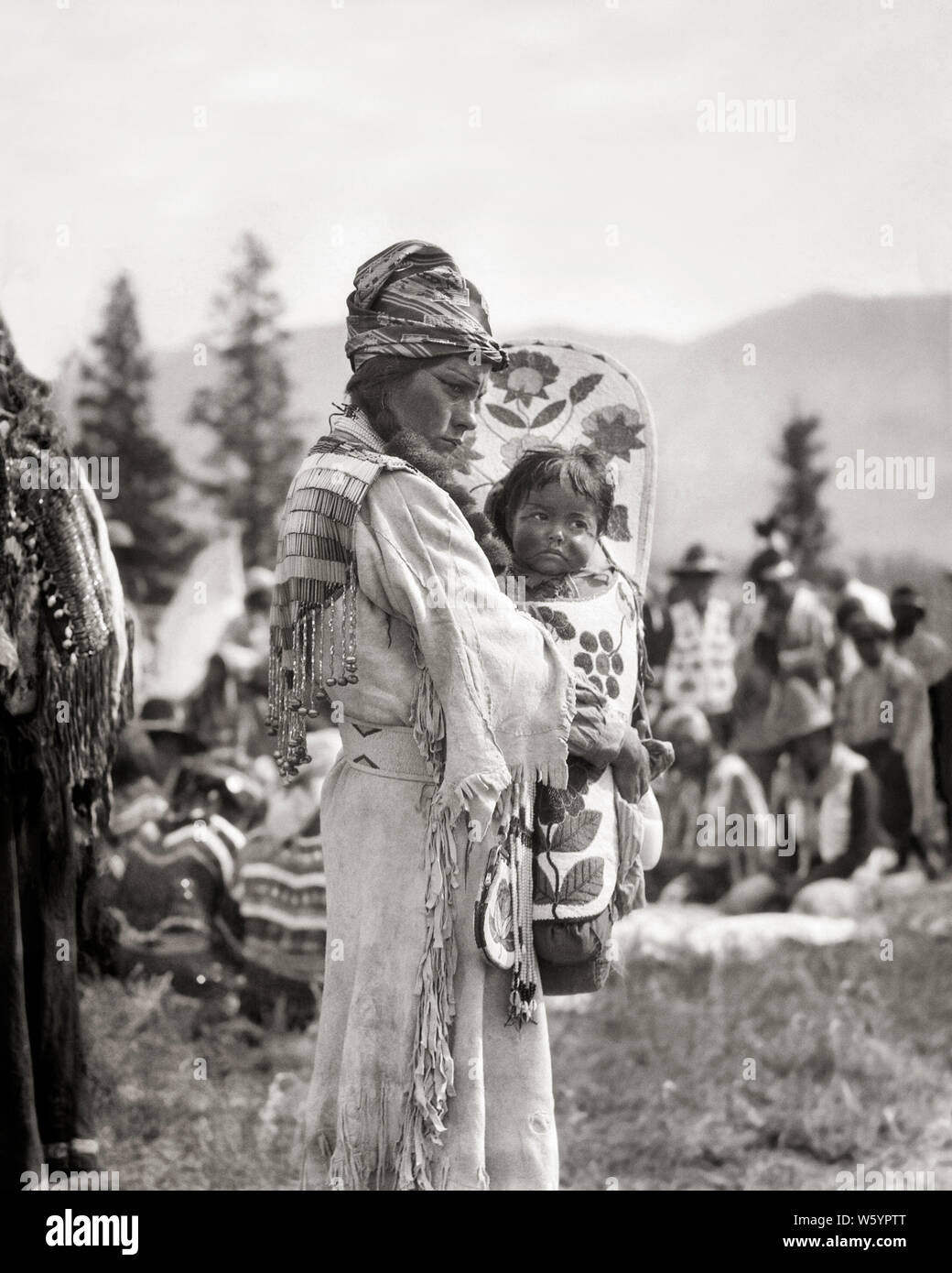 1920s 1930s NATIVE AMERICAN STONEY SIOUX INDIAN WOMAN MOTHER IN BEADED BUCKSKIN DRESS HOLDING BABY BOY GIRL IN PAPOOSE CANADA  - i59 HAR001 HARS FEMALES RURAL HOME LIFE COPY SPACE FULL-LENGTH HALF-LENGTH LADIES PERSONS TRADITIONAL INDIANS B&W NORTH AMERICA NORTH AMERICAN BUCKSKIN REAR VIEW IN CONCEPTUAL PAPOOSE NATIVE AMERICAN BABY BOY STONEY SIOUX BACK VIEW DEERSKIN FRINGED JUVENILES MID-ADULT MID-ADULT WOMAN MOMS NATIVE AMERICANS BABY GIRL BEADED BLACK AND WHITE CHILD CARRIER CRADLE BOARD HAR001 INDIGENOUS OLD FASHIONED Stock Photo