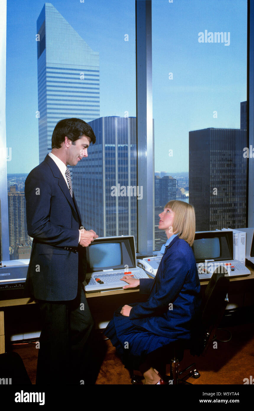 1980s URBAN OFFICE WITH EARLY HARDWARE WOMAN SEATED AT COMPUTER TERMINAL BY WINDOW TALKING TO MAN STANDING NEXT TO HER - ko1311 PHT001 HARS OLD FASHION 1 COMMUNICATION BLOND TECHNOLOGY TEAMWORK INFORMATION LIFESTYLE HISTORY FEMALES COMMUNICATING SEATED UNITED STATES COPY SPACE FRIENDSHIP FULL-LENGTH HALF-LENGTH LADIES COMPUTERS PERSONS UNITED STATES OF AMERICA MALES CONFIDENCE NEXT SCREENS BUSINESSWOMAN NORTH AMERICA NORTH AMERICAN SUIT AND TIE SELLING HIGH ANGLE EARLY NETWORKING MONITORS PROGRESS INNOVATION OPPORTUNITY AUTHORITY NYC OCCUPATIONS HIGH TECH CONNECTION ELECTRIC APPLIANCE HARDWARE Stock Photo