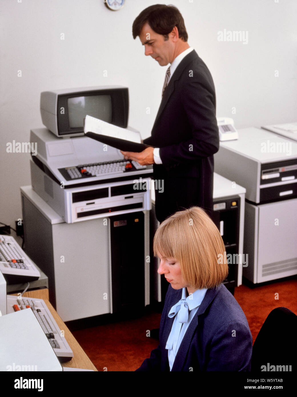 1980s OFFICE WITH EARLY COMPUTER HARDWARE WOMAN SEATED AT TERMINAL KEYBOARD MAN STANDING READING MANUAL - ko1320 PHT001 HARS KEYBOARD TECHNOLOGY TEAMWORK INFORMATION LIFESTYLE HISTORY FEMALES JOBS COMMUNICATING SEATED UNITED STATES COPY SPACE HALF-LENGTH LADIES COMPUTERS PERSONS MALES CONFIDENCE SCREENS BUSINESSWOMAN NORTH AMERICA WORK PLACE NORTH AMERICAN SKILL SUIT AND TIE OCCUPATION SELLING SKILLS HIGH ANGLE EARLY MANUAL NETWORKING MONITORS PROGRESS INNOVATION PRIDE OPPORTUNITY AUTHORITY OCCUPATIONS HIGH TECH CONNECTION ELECTRIC APPLIANCE HARDWARE STYLISH BUSINESSWOMEN TEXAS INSTRUMENTS Stock Photo