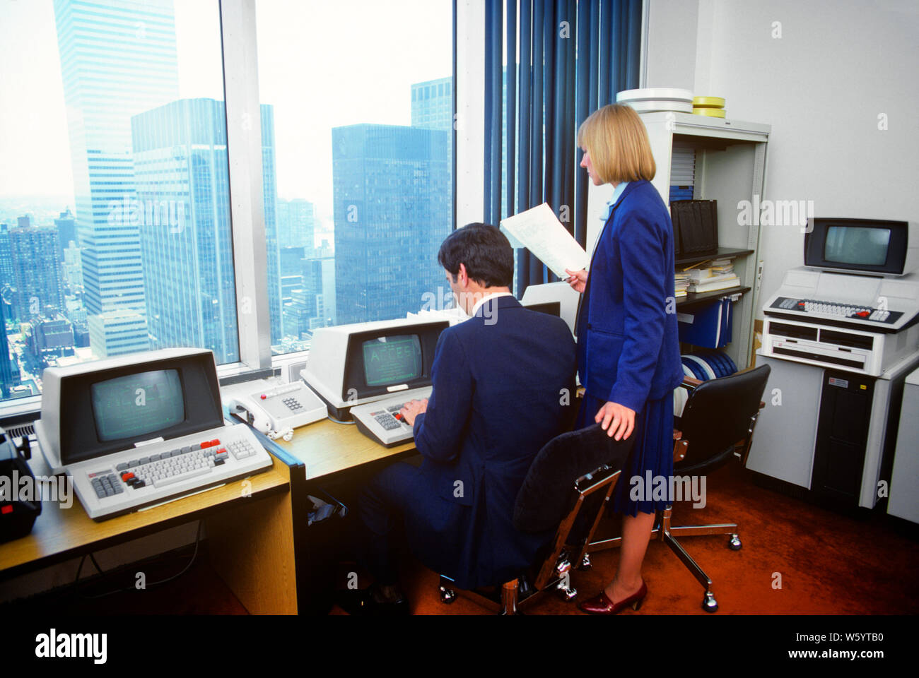 1980s URBAN OFFICE WITH EARLY HARDWARE MAN SEATED AT COMPUTER TERMINAL BY WINDOW WOMAN HOLDING PAPERS STANDING AT HIS SHOULDER - ko1310 PHT001 HARS INSTRUMENTS TECHNOLOGY TEAMWORK INFORMATION LIFESTYLE HISTORY FEMALES COMMUNICATING SEATED UNITED STATES COPY SPACE FULL-LENGTH HALF-LENGTH LADIES COMPUTERS PERSONS UNITED STATES OF AMERICA MALES CONFIDENCE SCREENS BUSINESSWOMAN NORTH AMERICA WORK PLACE NORTH AMERICAN SUIT AND TIE SELLING HIGH ANGLE EARLY HIS NETWORKING MONITORS PROGRESS INNOVATION PRIDE REAR VIEW OPPORTUNITY AUTHORITY OCCUPATIONS HIGH TECH CONNECTION ELECTRIC APPLIANCE HARDWARE Stock Photo