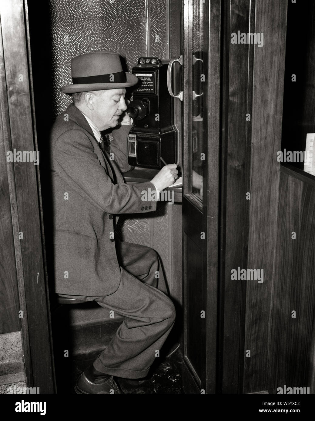 1940s MATURE MAN SITTING IN PAY TELEPHONE BOOTH TALKING ON PHONE AND TAKING WRITING NOTE NEW YORK CITY USA  - q40241 CPC001 HARS COMMUNICATING UNITED STATES COPY SPACE HALF-LENGTH PERSONS UNITED STATES OF AMERICA MALES MIDDLE-AGED B&W MIDDLE-AGED MAN SUIT AND TIE SELLING BOOTH DISCOVERY PAY AND OPPORTUNITY NYC OCCUPATIONS PHONES BOOKIE CONNECTION NEW YORK TELEPHONES CITIES STYLISH NEW YORK CITY COMMUNICATIONS COOPERATION SALESMEN BLACK AND WHITE CAUCASIAN ETHNICITY OLD FASHIONED Stock Photo