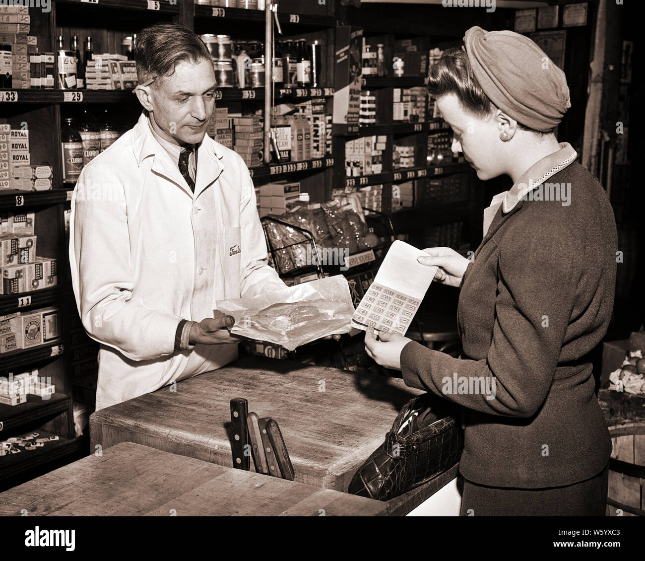 1940s WOMAN FOOD SHOPPER PURCHASING MEAT FROM BUTCHER WITH COUPON RATION STAMP BOOK IN GROCERY NEW YORK CITY USA  - q43052 CPC001 HARS MEAT BUTCHER B&W SERVICES SHOPPER SUCCESS GOODS CUSTOMER SERVICE CHOICE WORLD WARS WORLD WAR WORLD WAR TWO WORLD WAR II IN OCCUPATIONS CONCEPTUAL RATION RATIONING STYLISH SUPPORT WORLD WAR 2 NEW YORK CITY COUPON PURCHASING DEMAND MID-ADULT MID-ADULT MAN YOUNG ADULT WOMAN BLACK AND WHITE CAUCASIAN ETHNICITY OLD FASHIONED Stock Photo